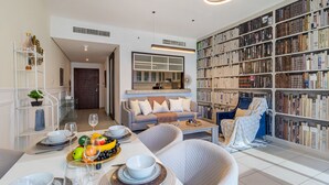A modern living room with a dining area, a bookshelf filled with books, and a kitchen visible through a glass door. - 2BR w Burj Khalifa View near Dubai Mall (Dubai)
