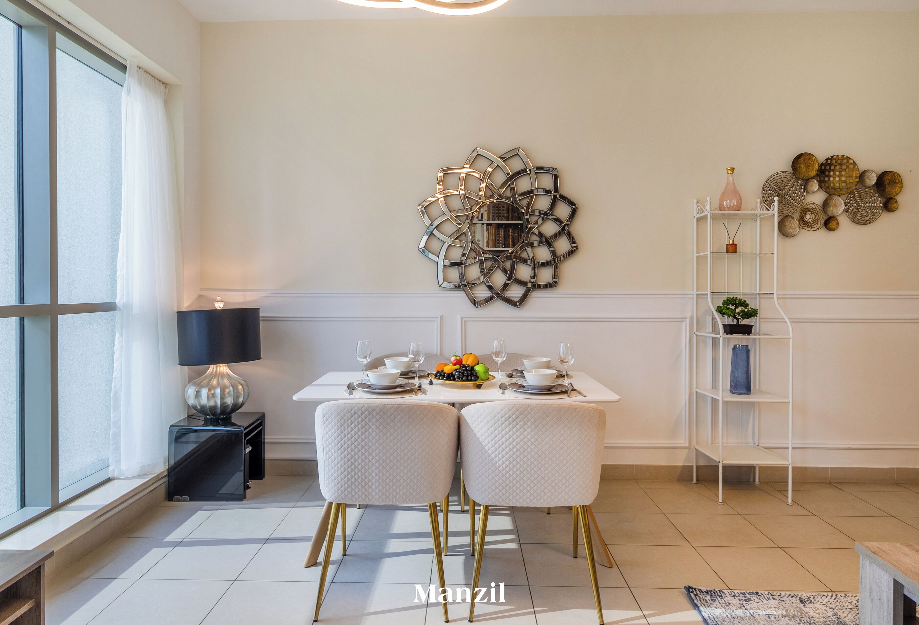 Et spiseområde med et hvidt bord, to hvide stole med gyldne ben, en dekorativ vægdekoration, en hvid reol med pynteting, og en sort lampe.