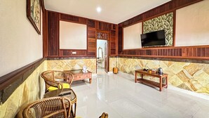 A spacious living area with a large flat-screen TV mounted on the wall, a wooden coffee table, wicker chairs, and a tiled floor. - Puri Karang Residence (Tembuku)