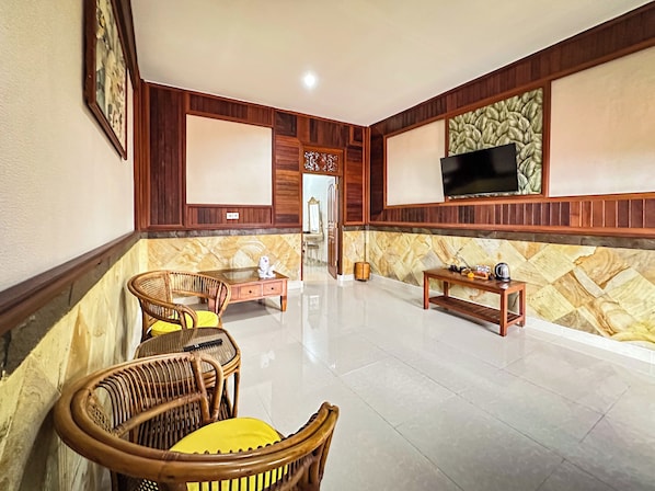 A spacious living area with a large flat-screen TV mounted on the wall, a wooden coffee table, wicker chairs, and a tiled floor. - Puri Karang Residence (Tembuku)