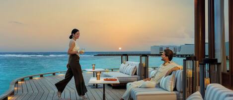 A couple on a deck with a sunset view, a pool, and a lounge area with cushions and tables.