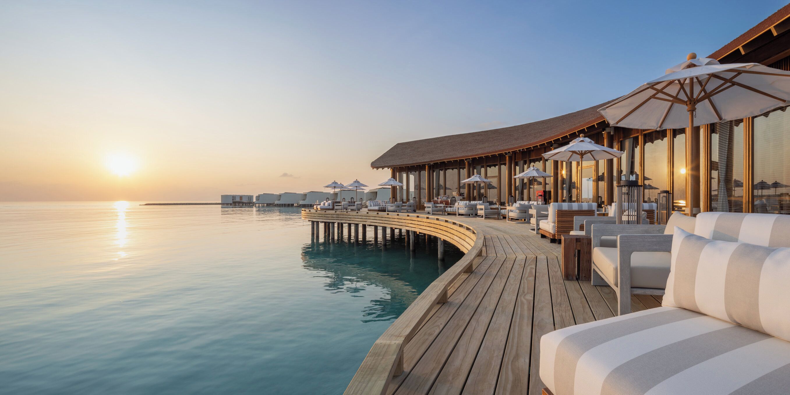 Eine Holzterrasse mit Liegestühlen und Sonnenschirmen mit Blick auf ein ruhiges Meer bei Sonnenuntergang.