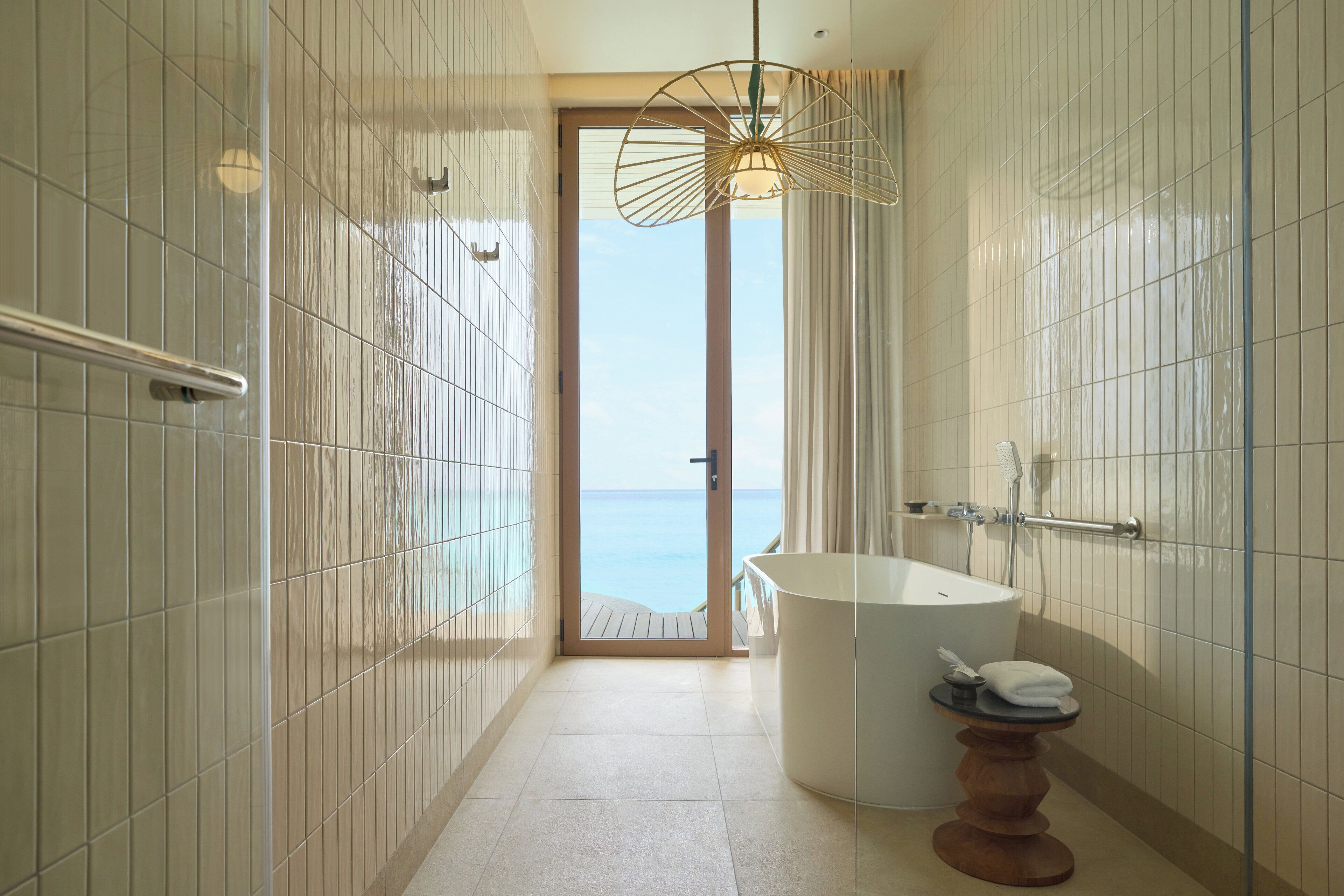 A modern bathroom with a large bathtub, a shower, and a view of the sea through an open door.