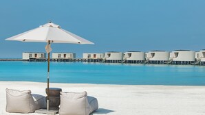 A beach with a white umbrella and two beige cushions, a clear blue sea, and overwater bungalows in the background. - Centara Grand Lagoon Maldives (Kodhipparu Falhu)