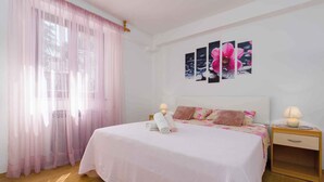 A bedroom with a bed, two bedside tables, a nightstand, a window with sheer curtains, and wall art.