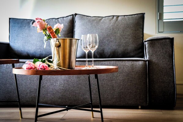 A small round table with a metal bucket and two wine glasses, a vase with pink flowers, and a dark grey sofa in the background. - The Wantage Suites (Johannesburg)