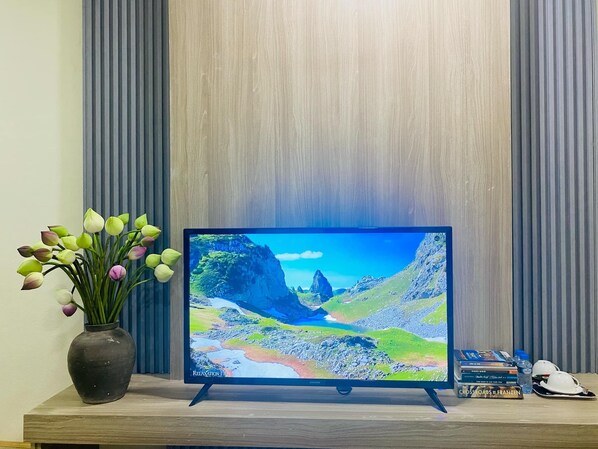 Un televisor de pantalla plana sobre un atril de madera muestra un paisaje pintoresco, un florero con flores y algunos libros, además de un casco sobre la mesa.