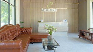 A reception area with a wooden reception desk, a brown leather sofa, a glass coffee table with a vase of flowers, and a large window providing natural light. - Peaceful Hotel (Vientiane)