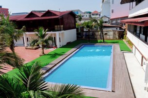 Una piscina rectangular con muelle de madera, rodeada por un área ajardinada con palmeras y un patio con baldosas rojas.