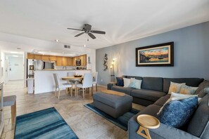 A modern living room with a grey sofa, a dining area with a white table and chairs, and a kitchen in the background. - Ohana Bliss l 2 Pools l Walk to Beach (St. Augustine Beach)