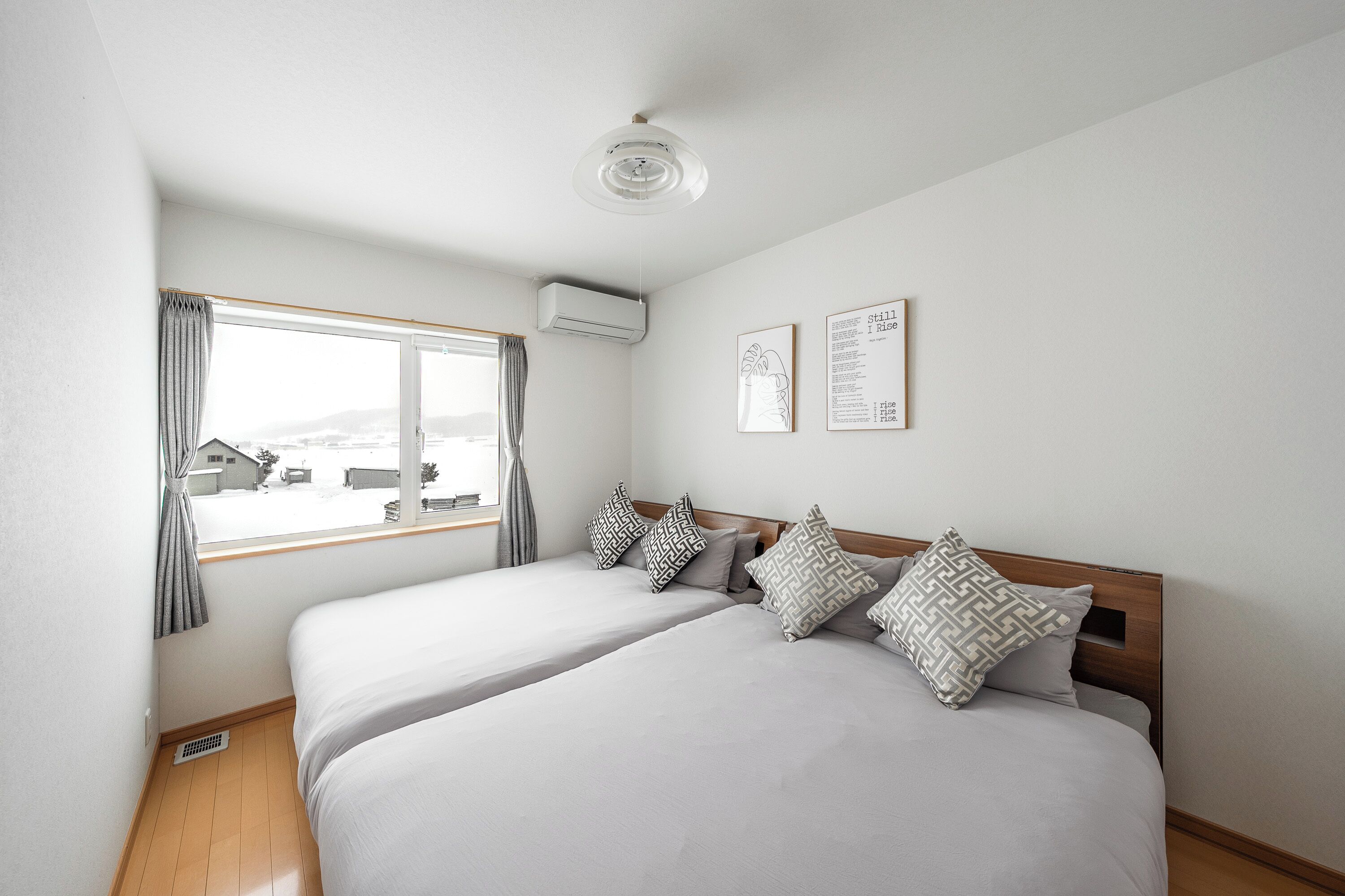 Een slaapkamer met twee bedden, een raam met gordijnen, een airconditioner en twee ingelijste foto's aan de muur.