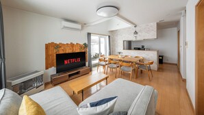 A modern living room with a flat-screen TV, wooden furniture, and a dining area with a brick backsplash. - CLEMATIS (Nakafurano)
