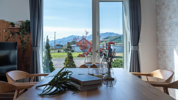 A dining area with a table set for two, a TV, and a view of mountains and a lake. - CLEMATIS (Nakafurano)