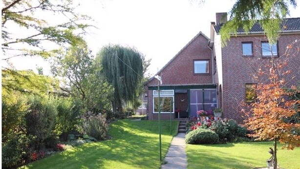 Une maison à deux étages, construite en briques, avec un jardin devant, un chemin menant à l’entrée et un petit potager où poussent diverses plantes et fleurs.