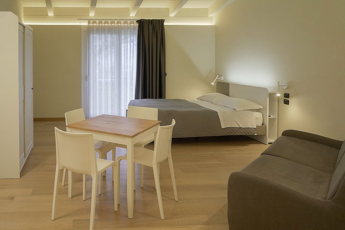 A hotel room with a bed, a desk with chairs, a sofa, and a window with curtains.