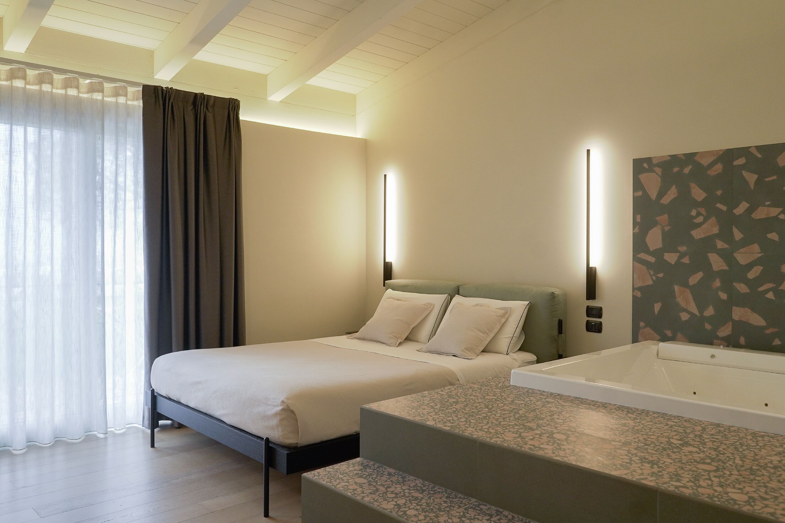 A hotel room with a bed, a small sofa, a bathtub, and a large window with curtains.