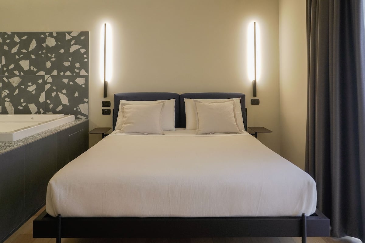 A modern hotel room with a large bed, two bedside tables, a built-in bathtub, and a wall-mounted light fixture.