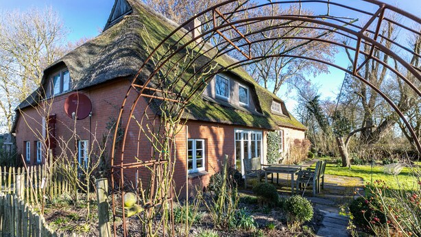 A house with a thatched roof, a brick exterior, and a garden with a wooden bench and table. - Holiday Apartment 'Reetdachhof Familie Dührsen' with Shared Terrace, Shared Garden and Wi-Fi (Norddeich)