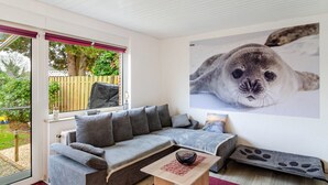 A modern living room with a grey sofa, a small wooden coffee table, and a large window with a view of greenery outside. - Bungalow 'Perle von Berumbur' with Private Terrace, Private Garden and Wi-Fi (Berumbur)