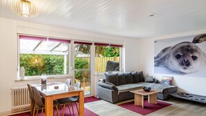 A modern living room with a dining table, sofa, and a large window with a view of greenery. - Bungalow 'Perle von Berumbur' with Private Terrace, Private Garden and Wi-Fi (Berumbur)