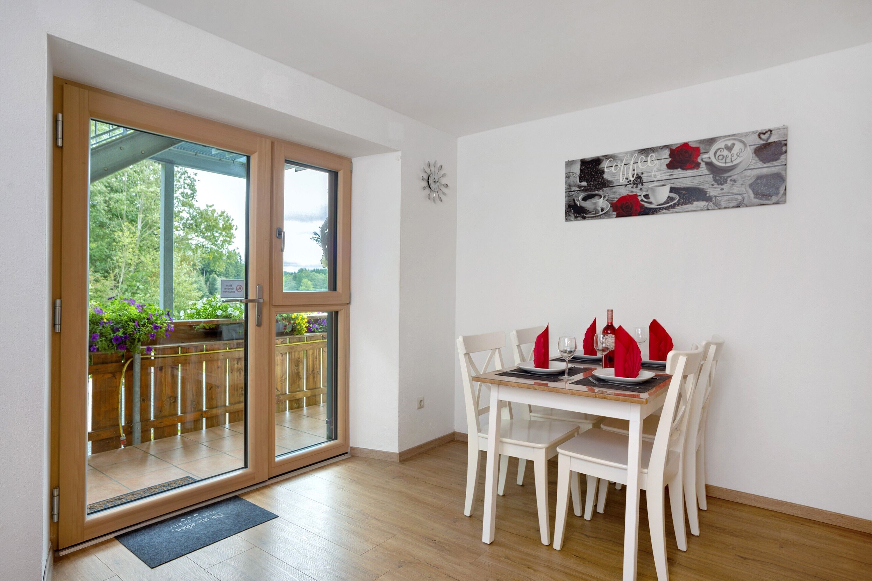 Een eetruimte met een tafel voor vier personen, witte stoelen, een houten vloer en een glazen deur die leidt naar een buitenruimte met een houten terras en groen.