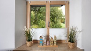Ein Fenster mit Blick auf Grünflächen, ein Regal mit kleinen Holzhausmodellen und zwei Blumentöpfe.