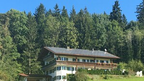 A multi-story building with a sloped roof, surrounded by a lush green field and dense forest.