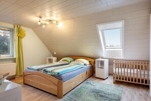 Une chambre avec un plafond en pente, un lit en bois, un berceau, une fenêtre et un radiateur.