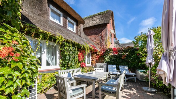 A patio with a table and chairs, surrounded by lush greenery and a thatched-roof building. - Apartment 'Fewo- Nommensen' with Shared Garden and Wi-Fi (Oland)