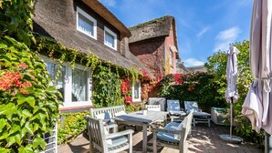 A patio with a table and chairs, surrounded by lush greenery and a thatched-roof building.