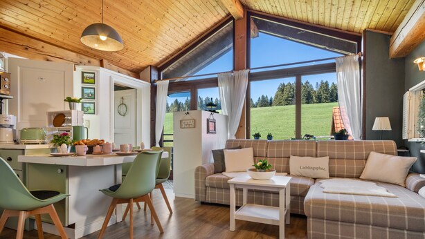 Ein gemütlicher Wohnbereich mit einer Couch, einem Esstisch und Stühlen, eine Küchenzeile mit Kühlschrank und Kaffeemaschine sowie große Fenster mit Blick auf eine grüne Landschaft.