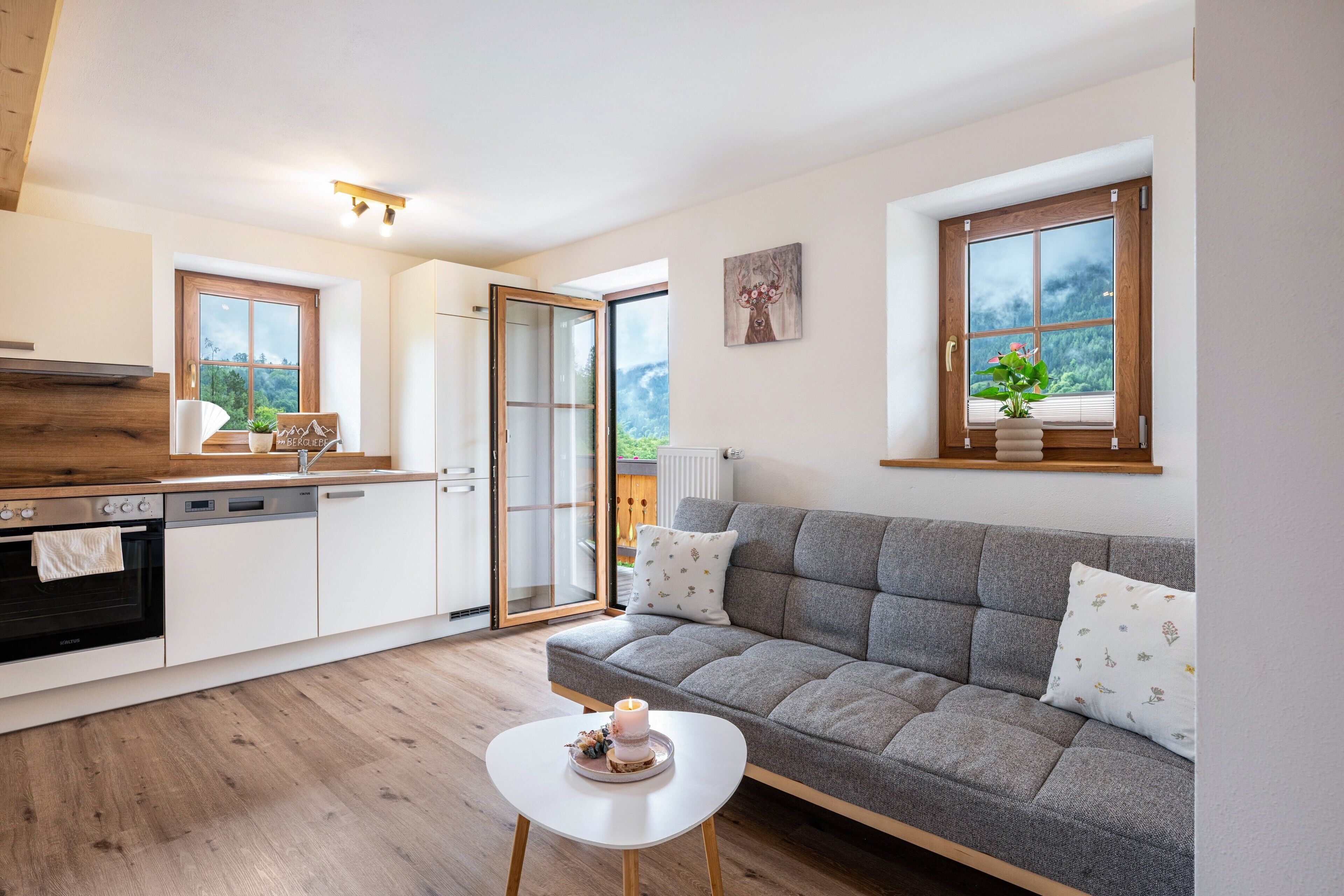 Een moderne keuken met een grijze bank, een witte tafeltje en uitzicht op de omgeving door een groot raam.