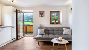 Un salon moderne avec un canapé gris, une petite table blanche et une vue sur un paysage verdoyant par une grande fenêtre.