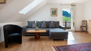 A modern living room with a sectional sofa, a black armchair, a wooden coffee table, and a view of a green landscape through a door. - Apartment 'Ferienwohnung Felix' with Balcony and Wi-Fi (Meßstetten)