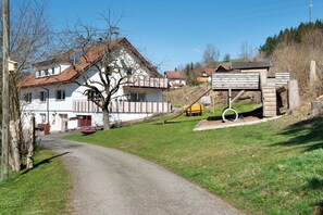 Un chemin de campagne mène à une maison blanche avec un toit rouge, entourée de verdure et d’une structure en bois dotée d’un toit en pente.