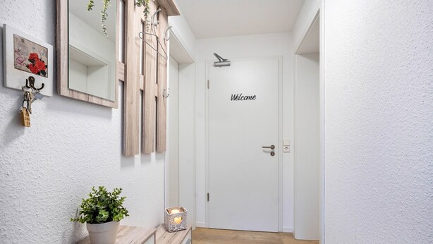Un pasillo con un banco de madera, una planta en maceta, un espejo en la pared y una puerta con un cartel que dice 'Welcome'.