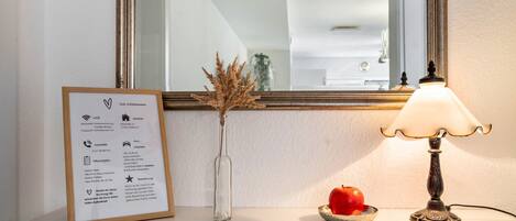 Un letrero de bienvenida enmarco, un florero con plantas secas, una manzana roja y una lámpara de mesa sobre una superficie blanca.