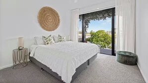 A bedroom with a large bed, a round woven wall decoration, a bedside table with a lamp, and a view through a sliding glass door. - Luxury Villas Allambi Broadbeach (With Car Parking) (Broadbeach Waters)