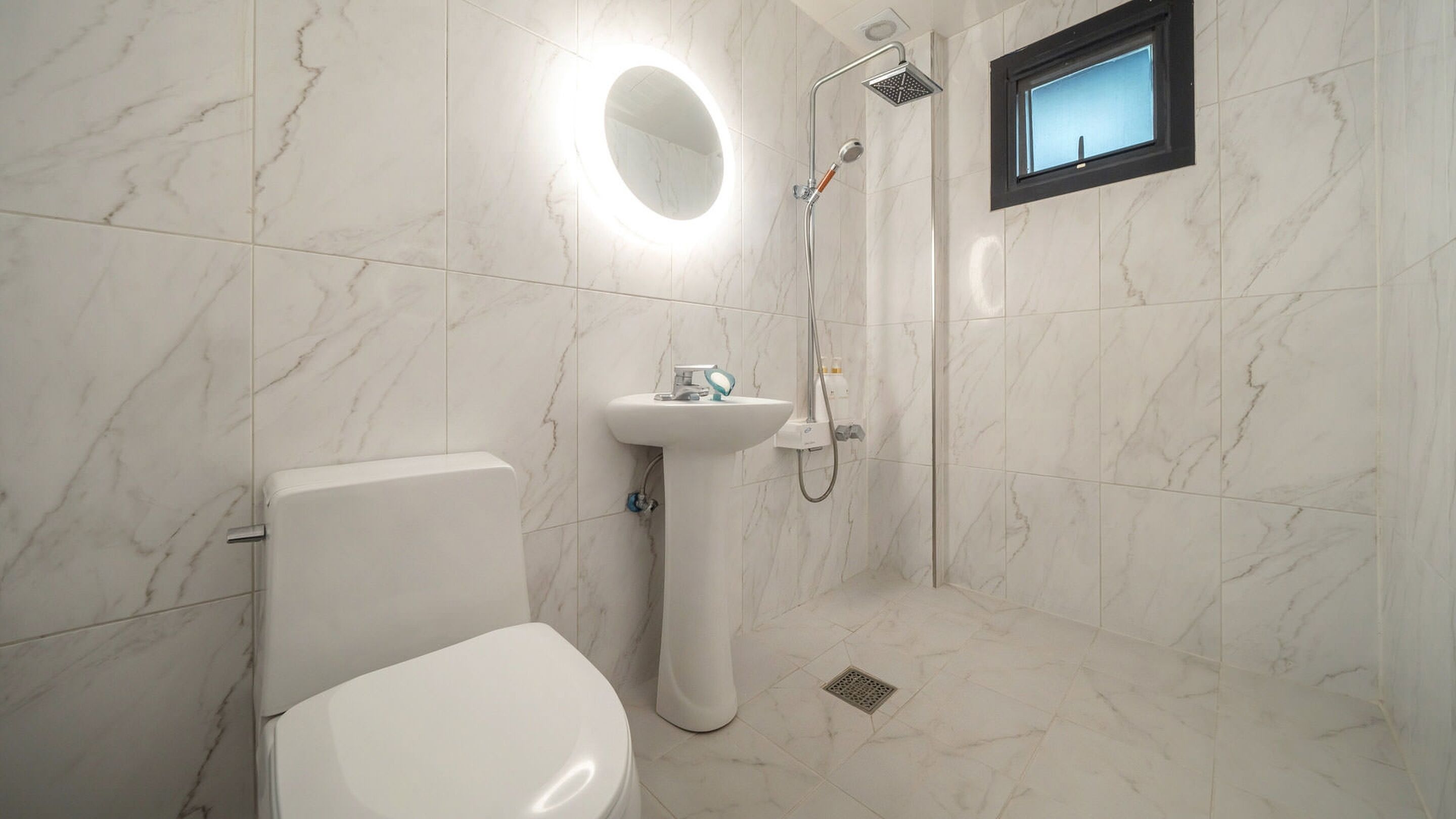A modern bathroom with a toilet, pedestal sink, shower, and a small window.