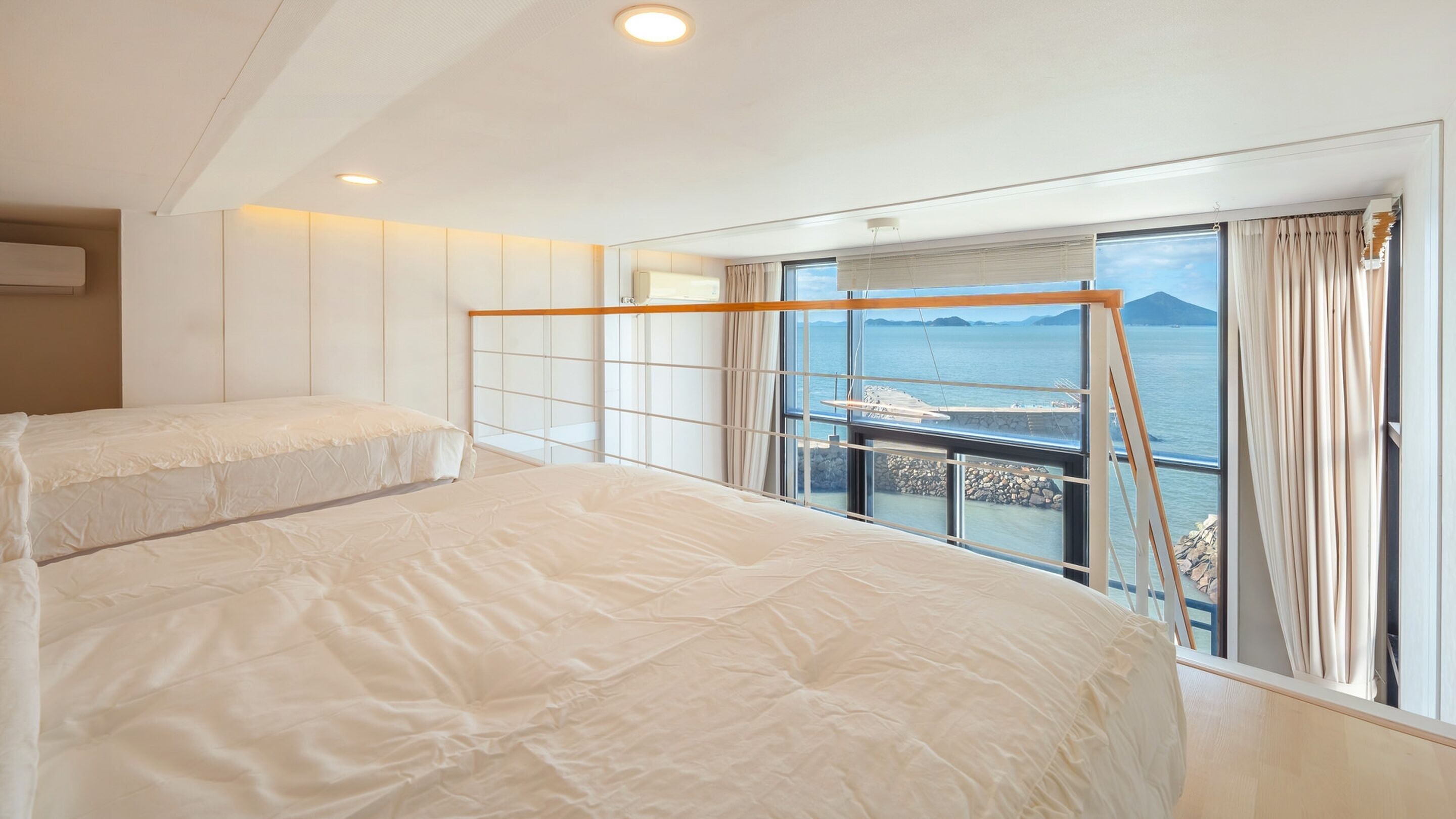 A modern hotel room with two beds, a large window with a view of the sea, and a balcony with a railing.