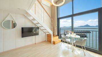 A modern living room with a TV, a staircase, a dining area with chairs, and a large window overlooking a body of water.