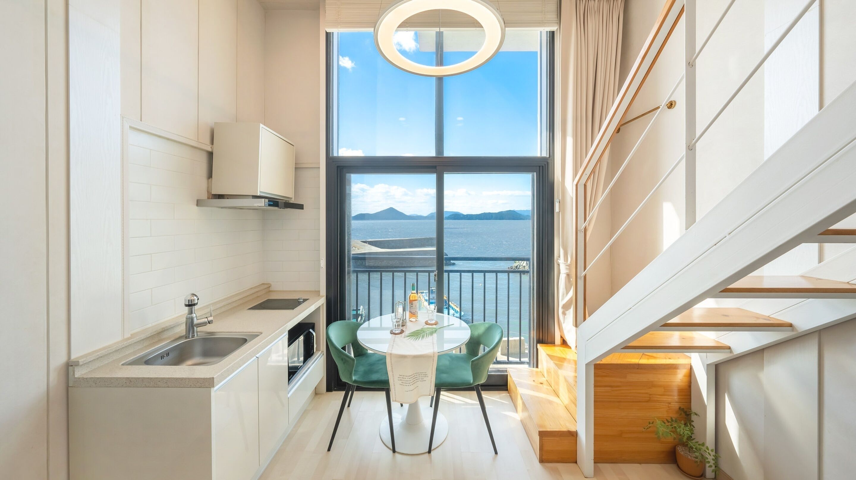 A modern kitchen with a view of the sea, a dining area with a table and chairs, and a staircase leading to an upper level.