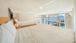 Habitación de hotel con una cama grande, cabecera de madera, mesita de noche con lámpara y vistas al mar a través de ventanas amplias.