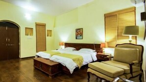 Een hotelkamer met een tweepersoonsbed, een houten bankje, een stoel, een nachtkastje met een lamp en een deur.