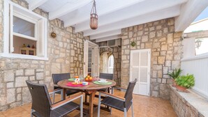 Um pátio com paredes de pedra e uma mesa de madeira redonda posta para quatro pessoas, rodeada por cadeiras de vime.