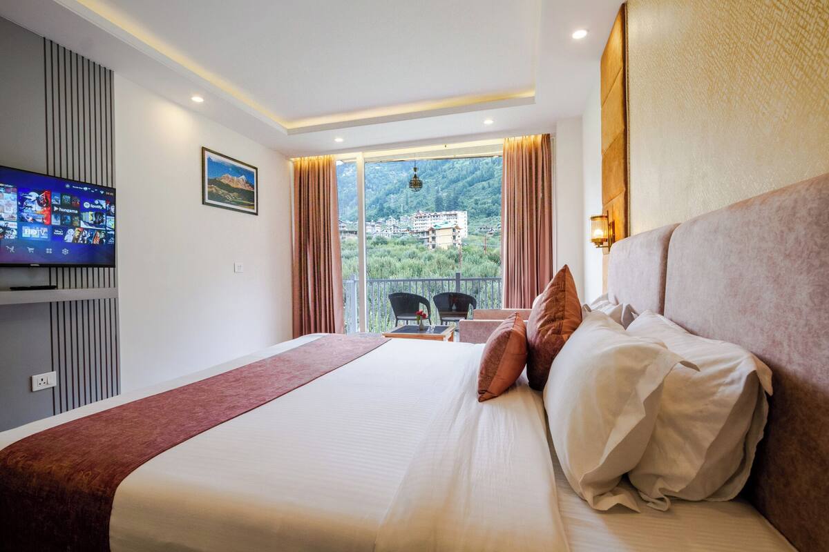 Una habitación de hotel con una cama grande, un televisor en la pared, un balcón con vistas a las montañas y una mesa con sillas.