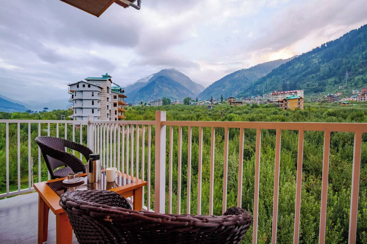 Un balcón con mobiliario de mimbre, una mesa y vistas a un paisaje montañoso con edificios al fondo.