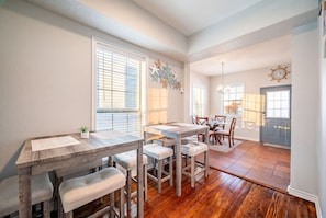 A dining area with a wooden table and white upholstered chairs, a kitchen in the background, and a large window with blinds.