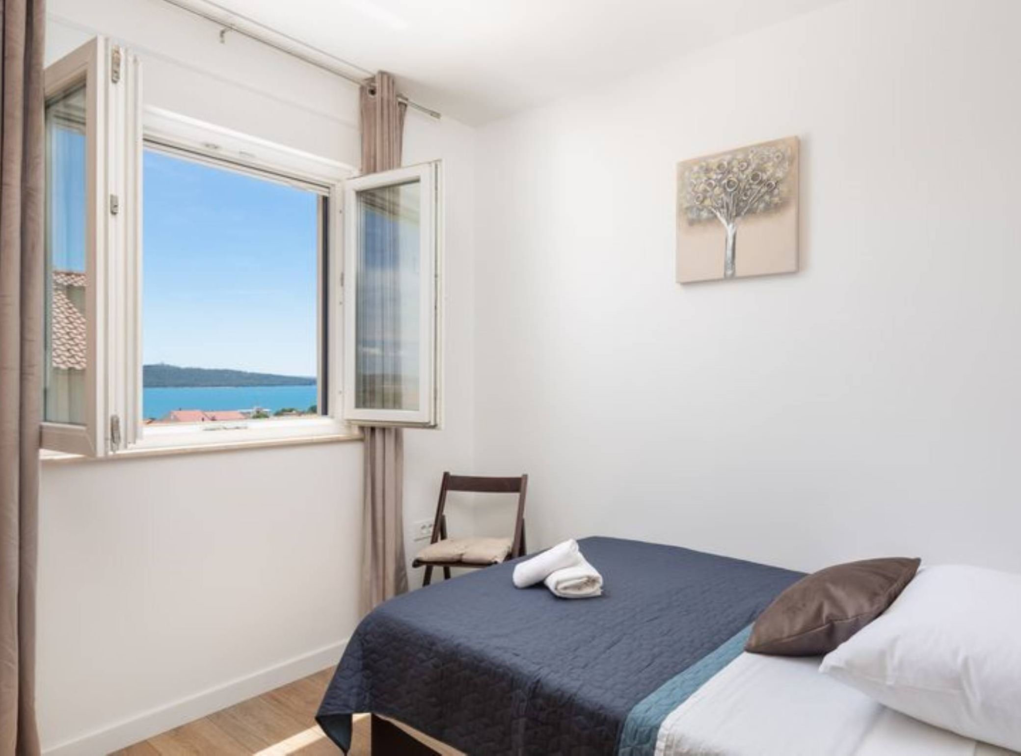 Une chambre à coucher comprenant un lit, une chaise, une fenêtre donnant sur la mer et une œuvre d’art murale.