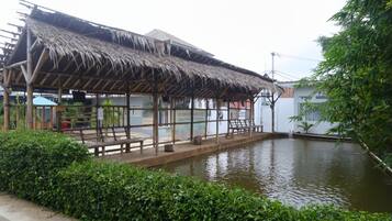 Ein traditionelles Gebäude mit einem Reetdach, Bänken und einem kleinen Pool, umgeben von viel Grün.